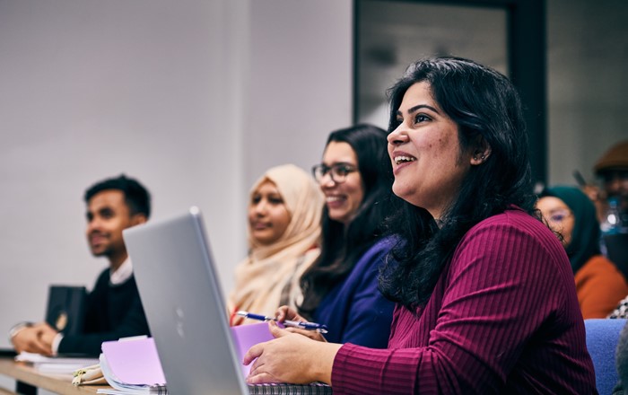 A group of people sit in a classroom setting, with one woman in the foreground using a laptop. 