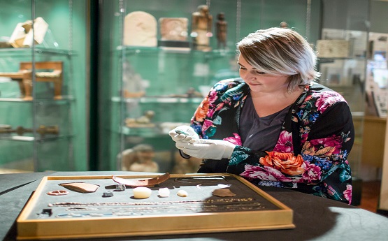 A blonde woman wearing a colourful, flowery top is stood next to glass display cabinets looking at items from the Egypt Collection.