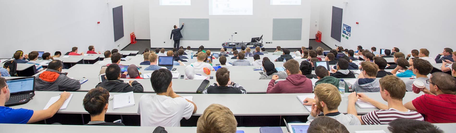 A view from the back of a lecture theatre of students sat in rows and a lecturer pointing at the board at the front of the class