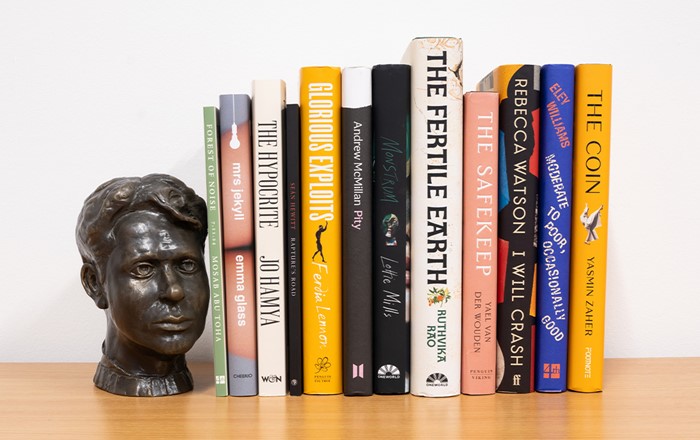 A small bust of Dylan Thomas and 12 books on a wooden shelf against a white background.