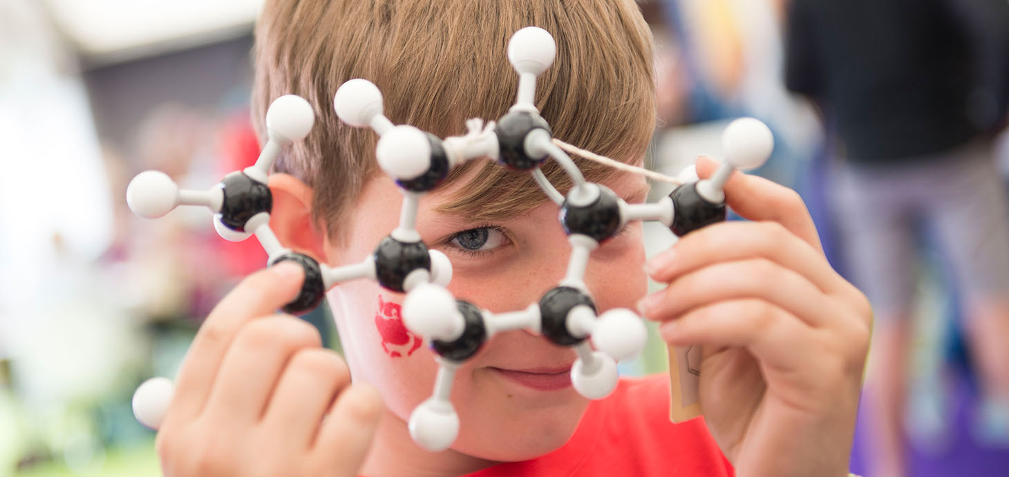 A child holds a molecular model made of black and white balls and sticks close to their face, peering through it with one eye.