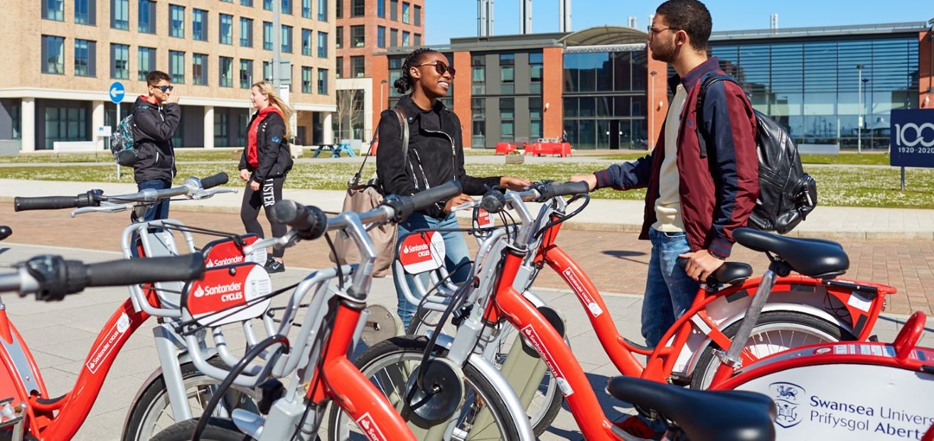 Four students standing outdoors in front of buildings by bicycles.