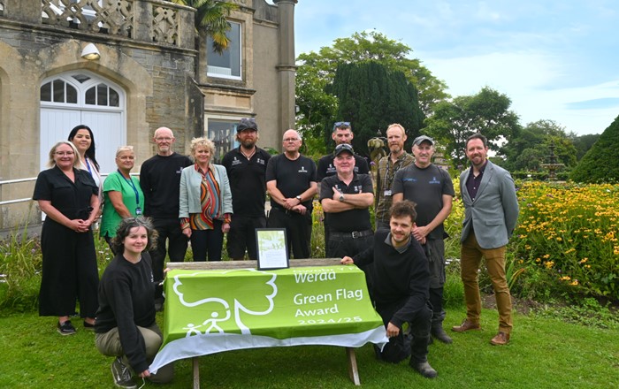 Group of people standing in a garden standing outside a building by a table draped with a green flag.