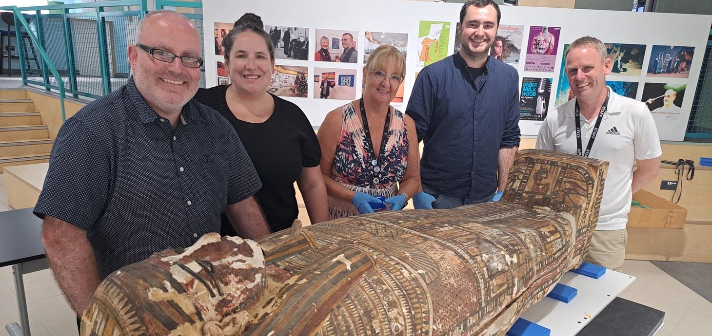 Five people in a gallery standing behind an Egyptian coffin.