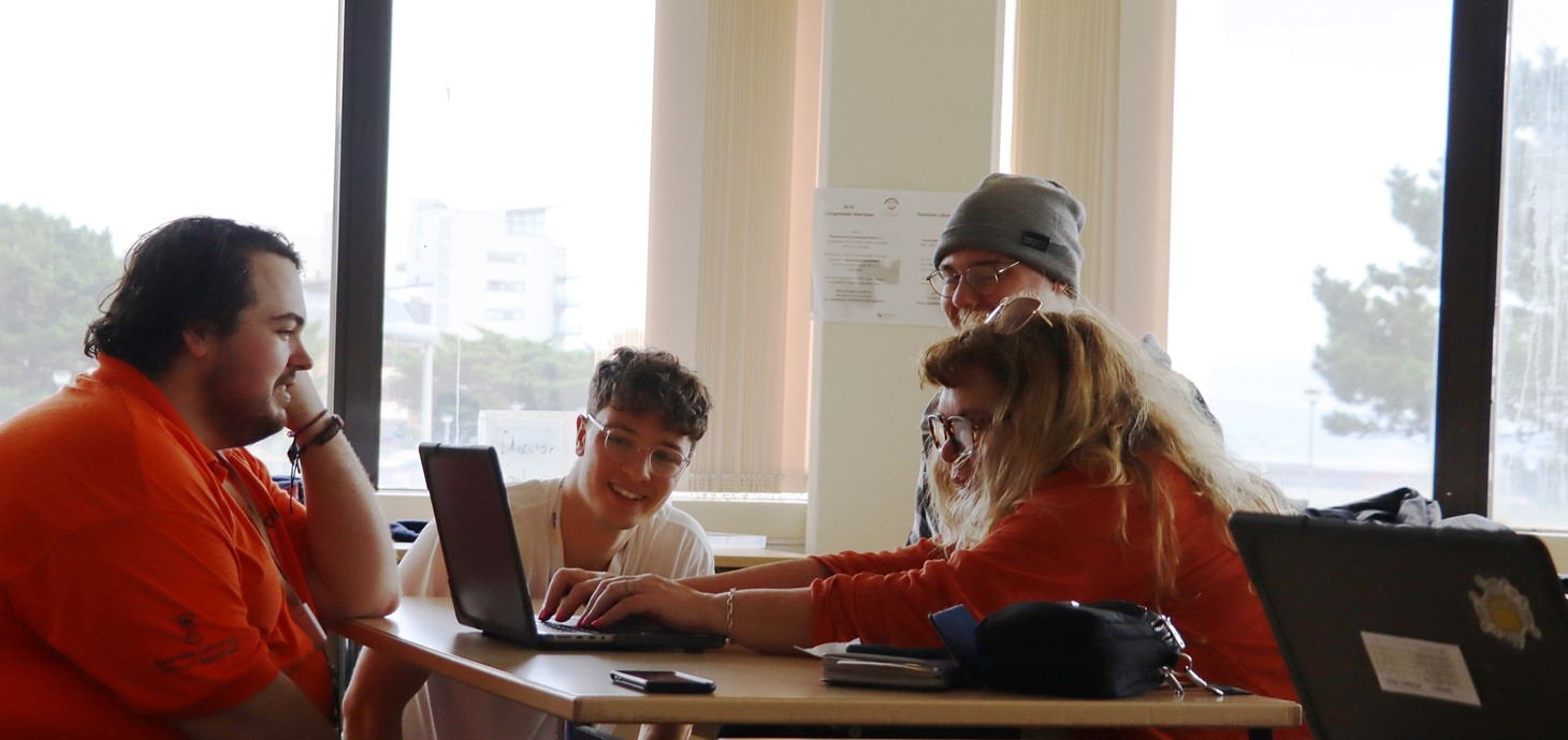 Four people are at desks in a classroom looking at laptop screens. 