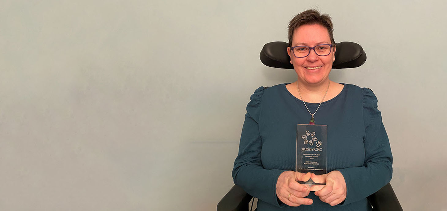 Alt-text Dr Aimee Grant is smiling as she holds her glass Autism CRC award. She is seated in her wheelchair. 