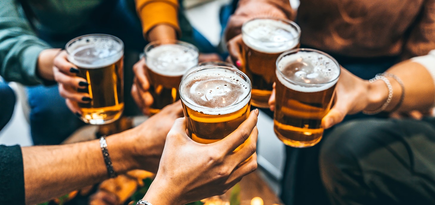 Shot of a group of people's hands. They are all holding full pint glasses of beer above a table top and clinking them together to raise a toast.