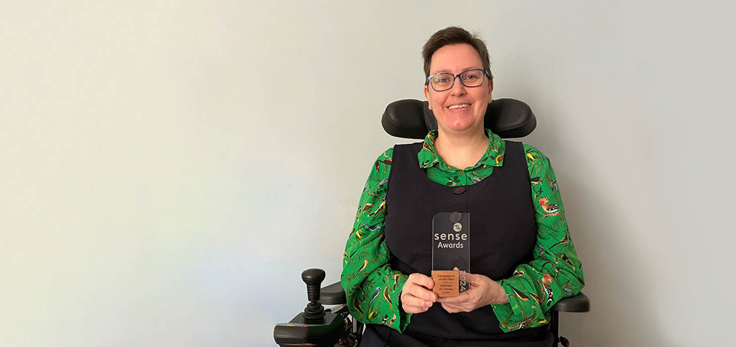 Dr Aimee Grant is smiling as she holds her glass Sense award. She is seated in her wheelchair and wears a black dress and green blouse.