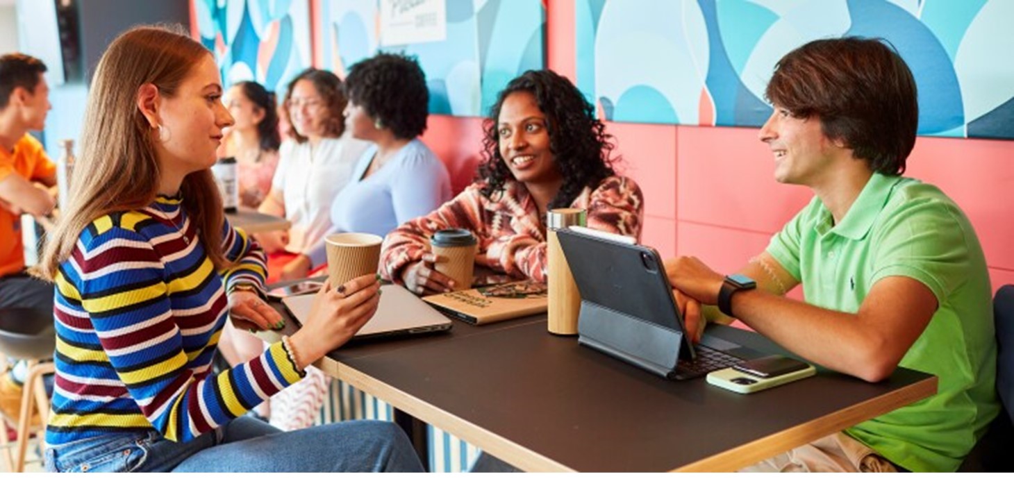 Three students are in lively conversation in a brightly coloured coffee shop. They have laptops, tablets and phones and are holding drinks cups.