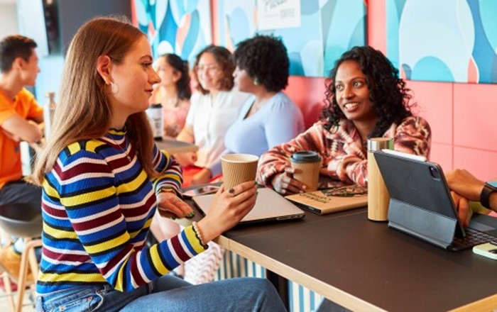 Three students are in lively conversation in a brightly coloured coffee shop. They have laptops, tablets and phones and are holding drinks cups.