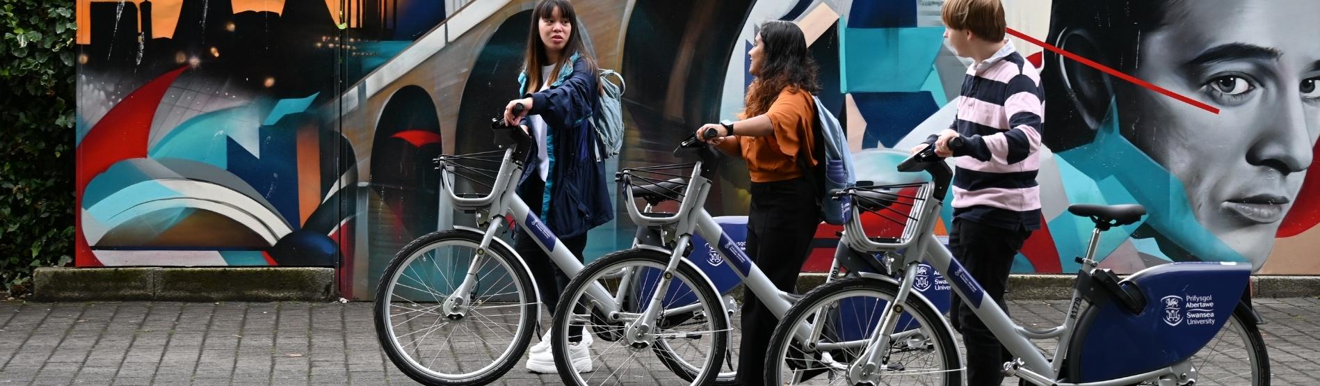 Students walking with Swansea University Cycles bikes on campus 